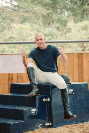 Man sitting on a blue equestrian obstacle with two thumbs up, wearing riding boots and a dark shirt.