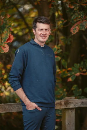 Man wearing a blue sweater standing outdoors with trees and foliage in the background