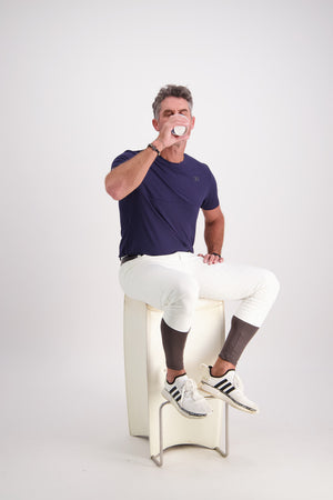Man sitting on a white stool wearing a navy blue t-shirt, white pants, and black Adidas shoes.