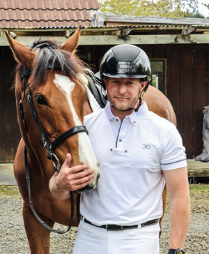 Man wearing a white mens show shirt standing next to a brown horse outdoors