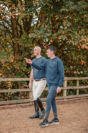 Two men in equestrian attire standing in a field with trees in the background