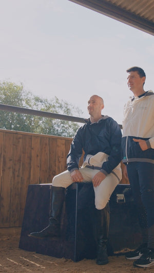 Two men in equestrian attire standing on a wooden platform with a clear sky background.