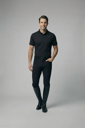 Man wearing black mens polo shirt and equestrian pants with boots standing against plain background