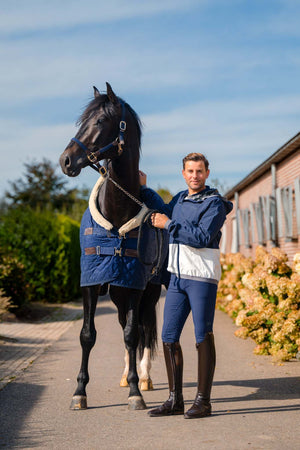 Man in equestrian attire standing next to a horse wearing a blue rug on a path with buildings and trees in the background.